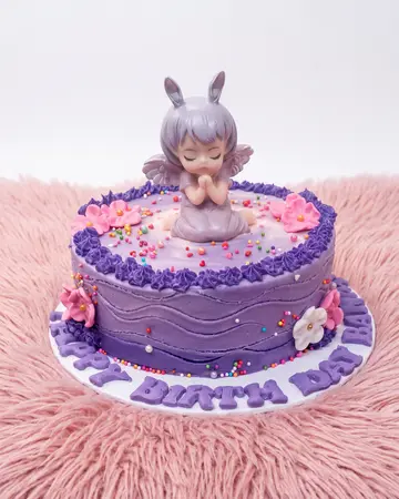 A lilac cake with piped shell borders, sugar blossoms and a seated fairy figure on top.